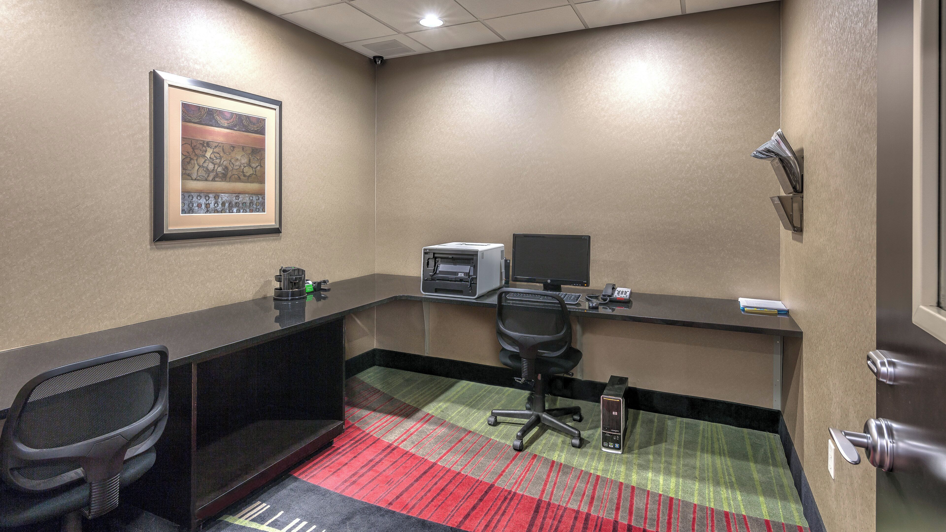 A hotel room with a desk, chair, printer, and a framed picture on the wall.