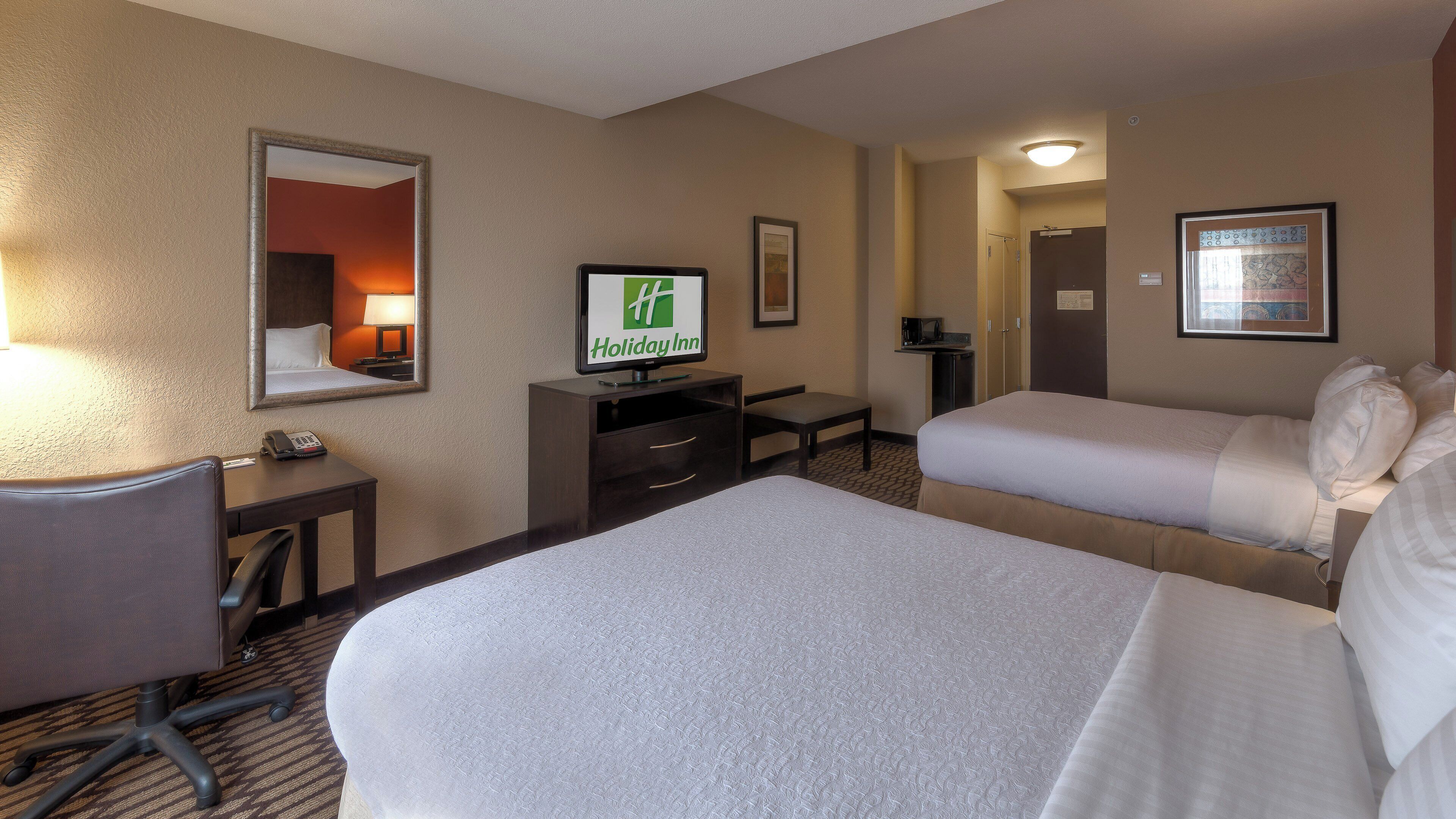 A hotel room with two beds, a desk with a computer, a telephone, and a television displaying the Holiday Inn logo.