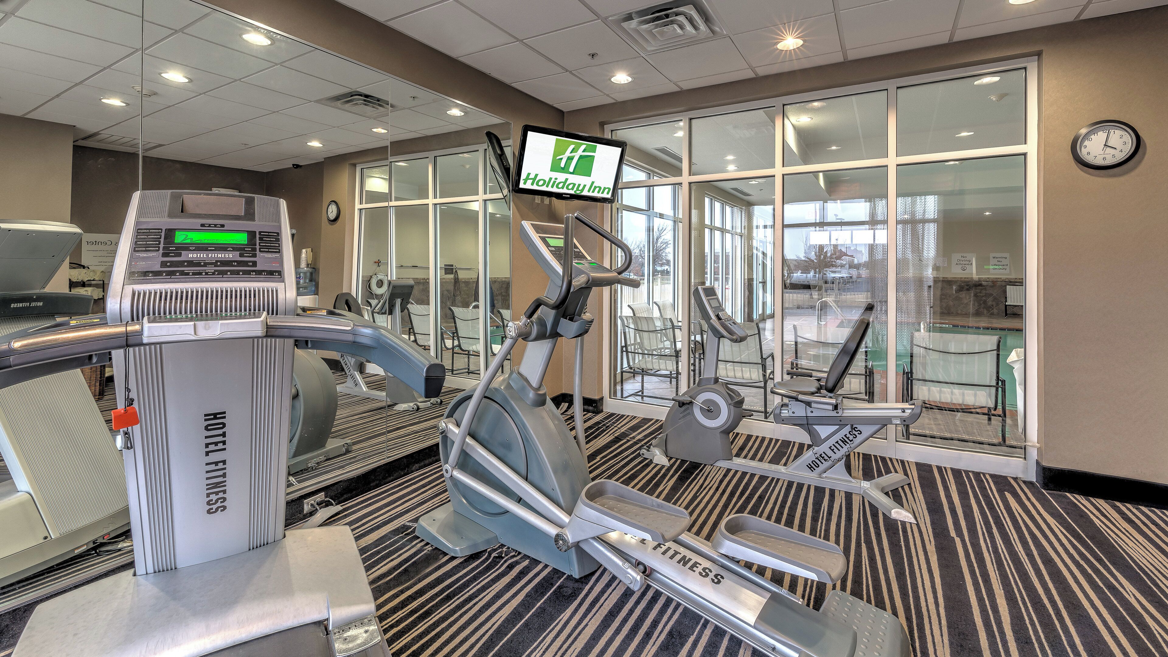 A hotel fitness center with exercise equipment, a digital display, and a Holiday Inn sign.