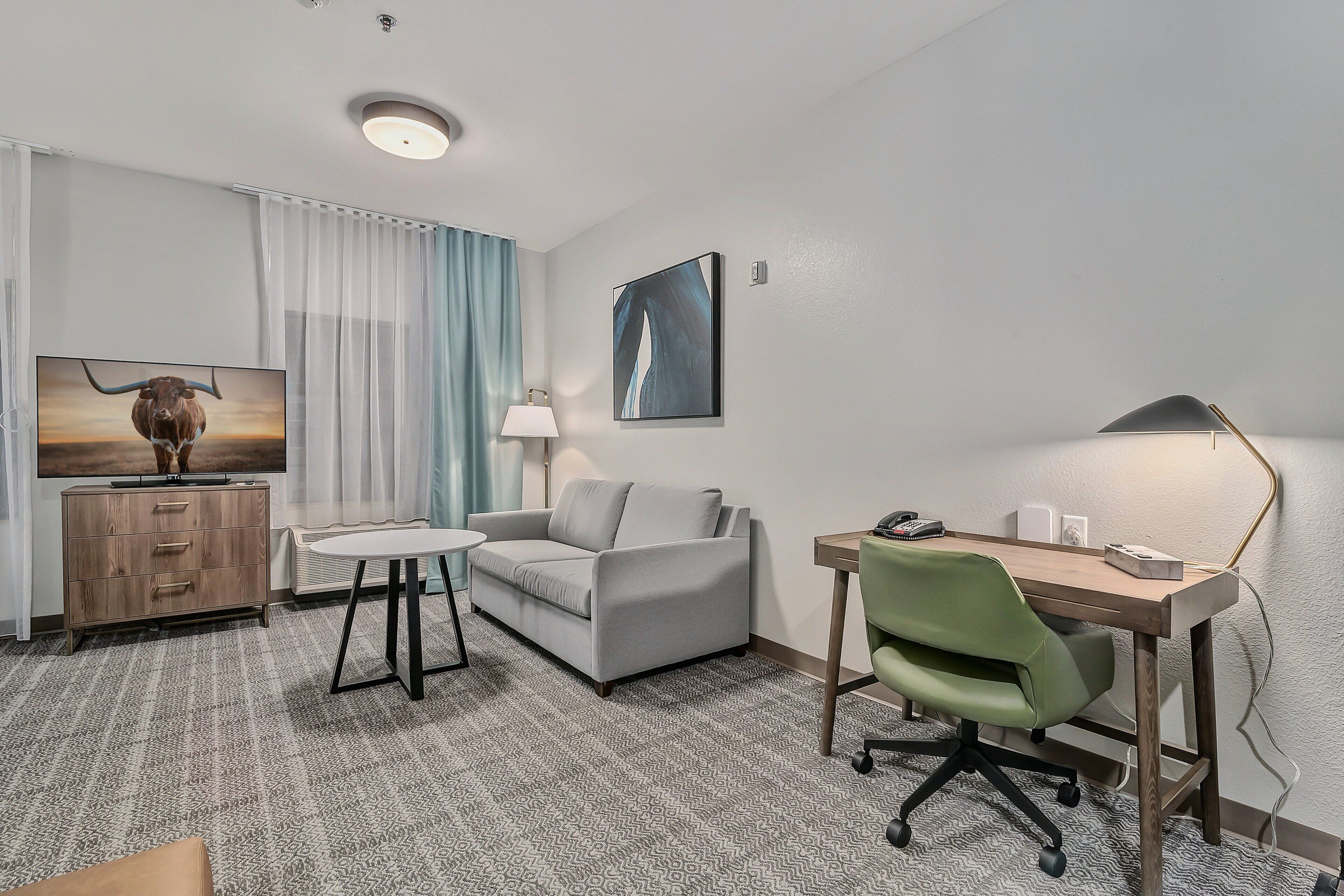 A hotel room with a sofa, a desk with a chair, a television, a lamp, and a painting on the wall.