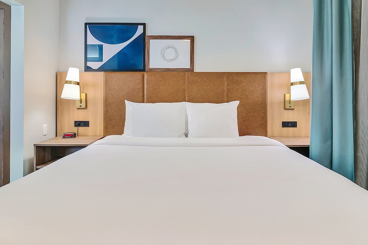 Habitación de hotel con una cama grande, dos mesitas de noche con lámparas, un reloj y arte abstracto en la pared.