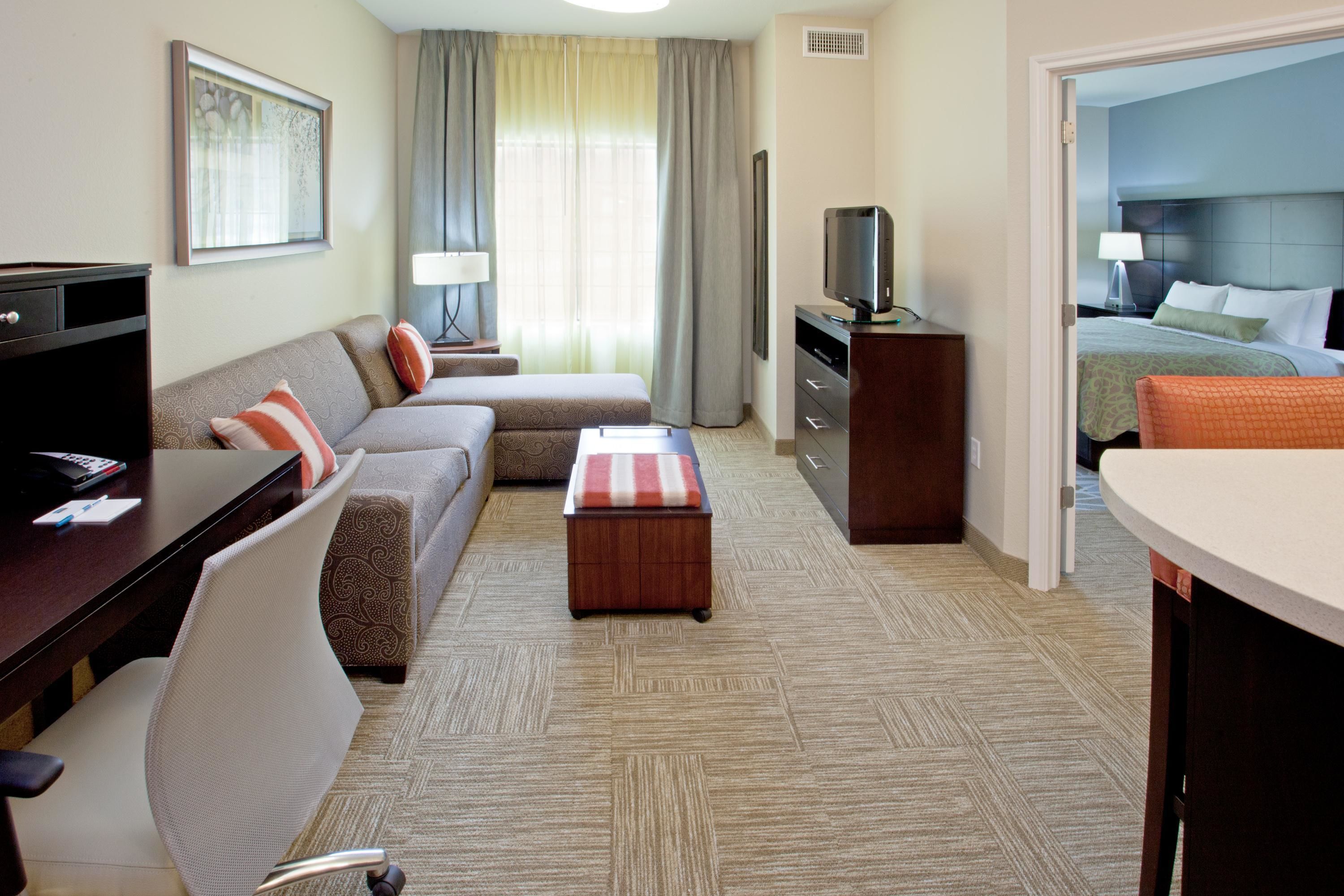 A hotel room with a sofa, a desk, a television, a small ottoman, and a bed with a headboard.