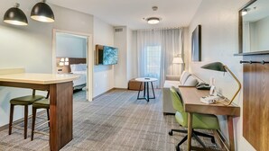Ein Hotelzimmer mit Schreibtisch aus Holz, zwei Stühlen, einem kleinen runden Tisch, einer Couch, einem an der Wand befestigten Fernseher und einem großen Spiegel.