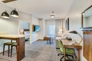 A hotel room with a wooden desk, two chairs, a small round table, a sofa, a TV mounted on the wall, and a large mirror. - Staybridge Suites University Area by IHG (Tyler)