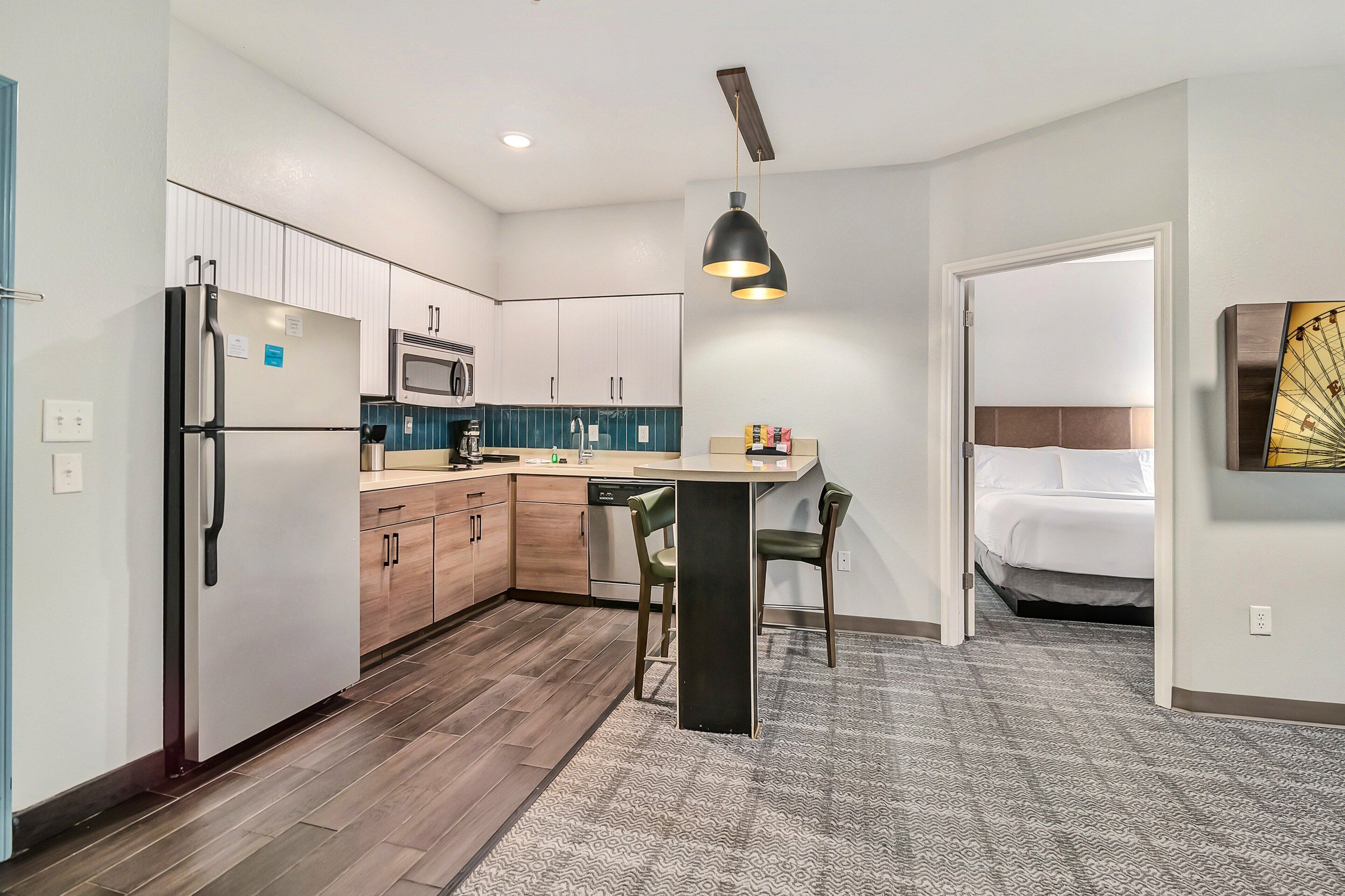 A compact kitchen with modern appliances, a dining area with a bar stool, and a bedroom with a bed and a wall-mounted artwork.