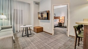 Ein Hotelzimmer mit einer Couch, einem kleinen Tisch, einem braunen Hocker, einem Gemälde eines Stiers und Blick in einen weiteren Raum mit einem Bett und einem Nachttisch.