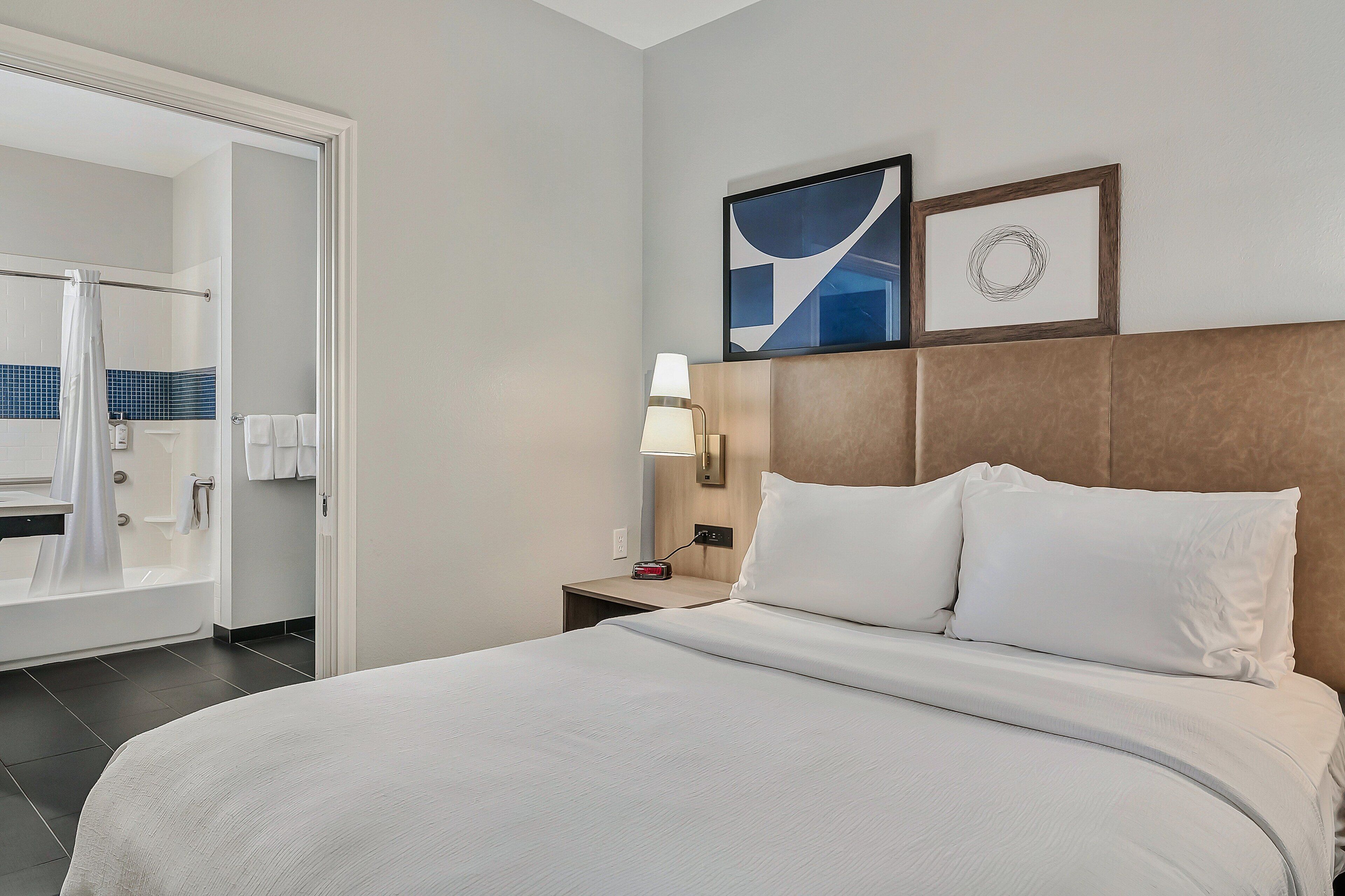 A hotel room with a bed, a nightstand with a lamp, a bathroom with a shower, and a wall with two framed pictures.