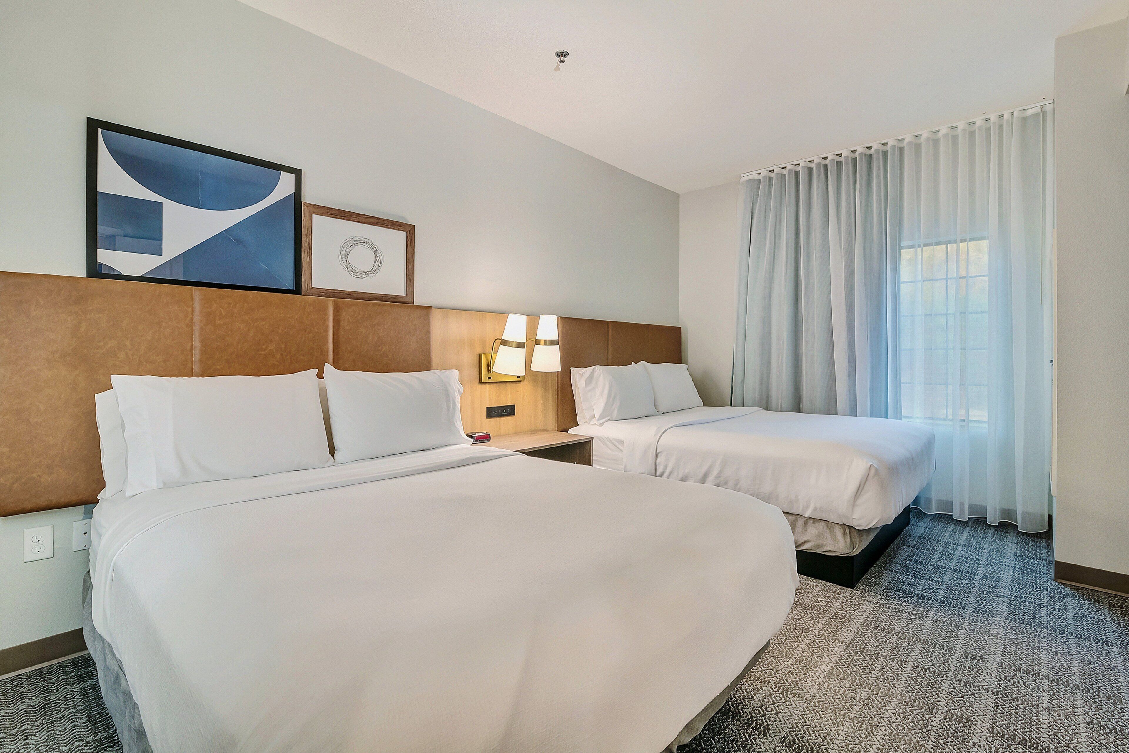 A hotel room with two beds, a headboard, a bedside table with lamps, a window with curtains, and a wall with framed artwork.
