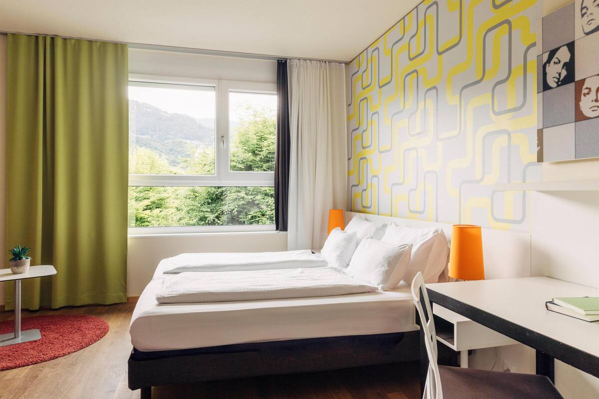 Ein Hotelzimmer mit Bett, Schreibtisch, Stuhl, einem Fenster mit Vorhängen und Blick auf Grünfläche.