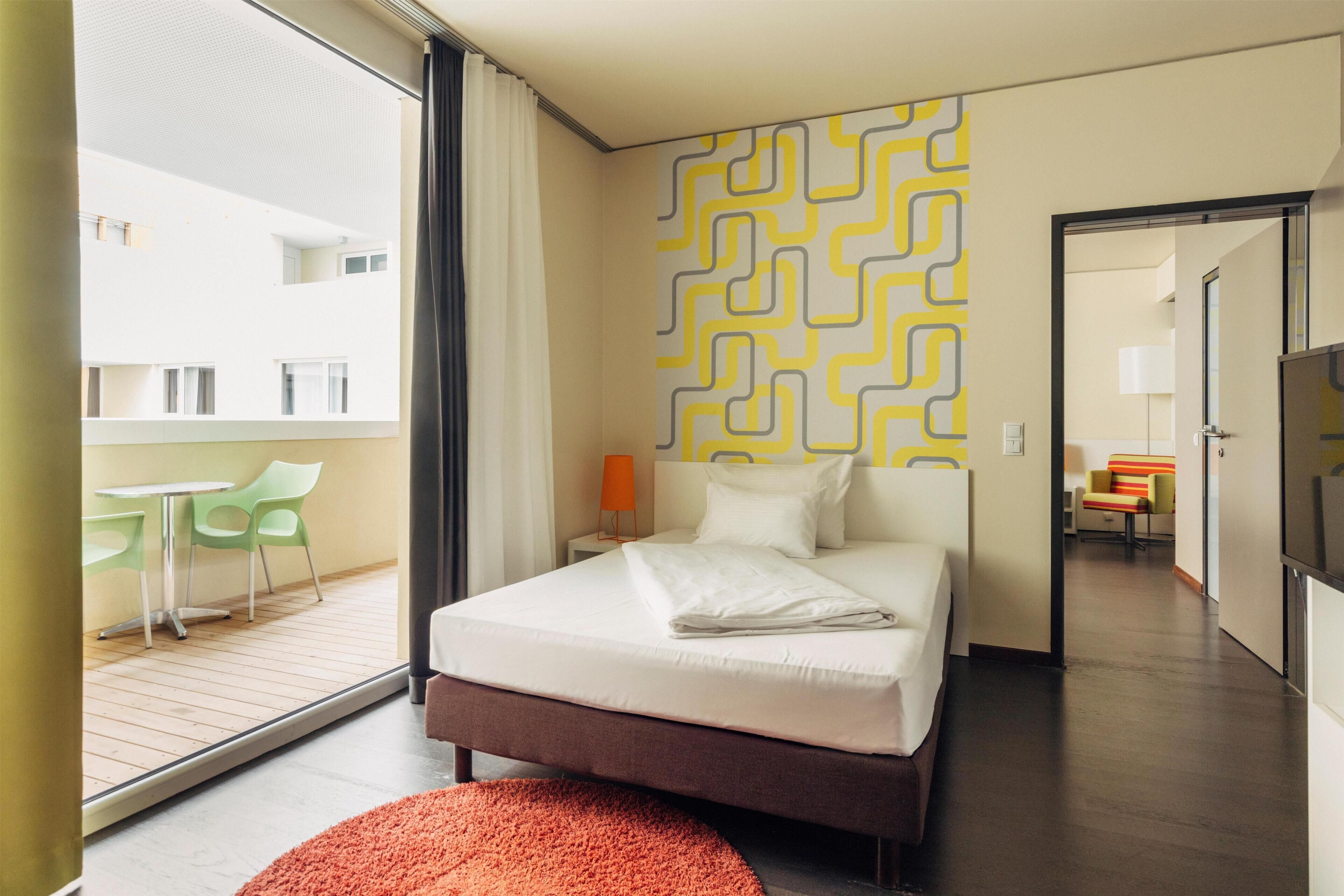 A modern hotel room with a large bed, a view to the outside, a patterned wall, and a small table with chairs.