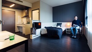 Een moderne hotelkamer met een bed, nachtkastje en een persoon in een rolstoel.