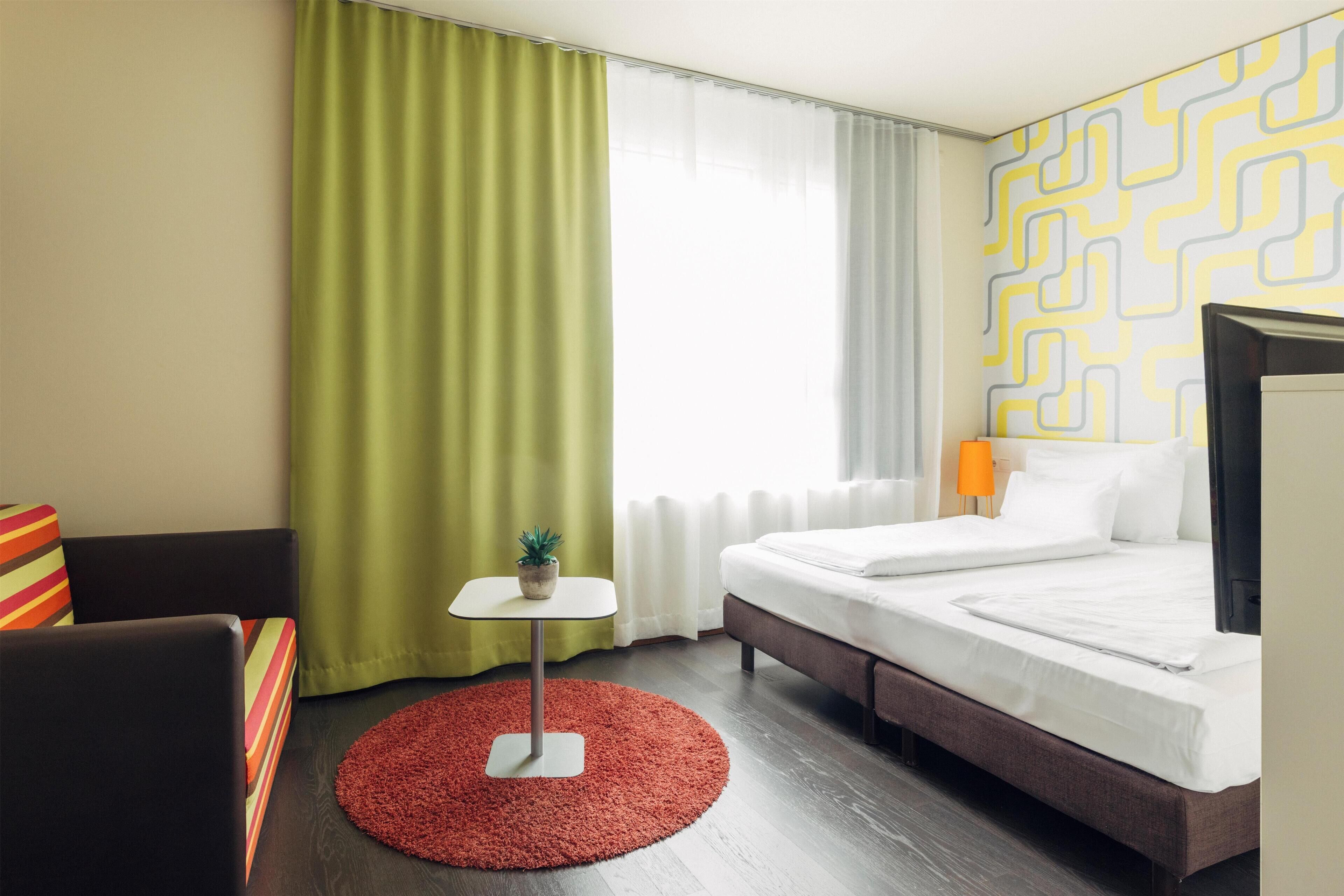 Ein Hotelzimmer mit einem Bett, einem kleinen Tisch mit einer Pflanze, einem Sessel und einem roten Teppich.