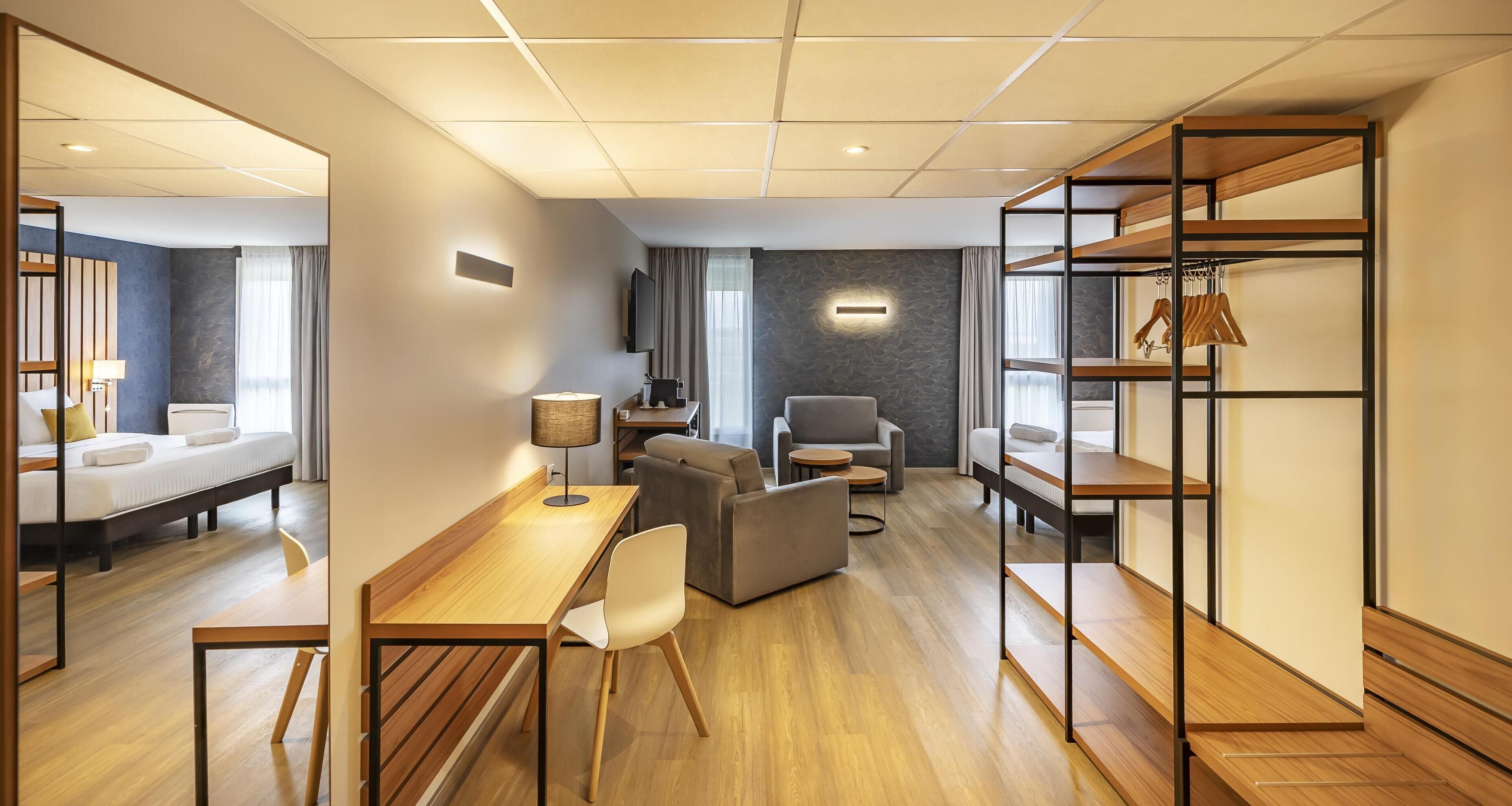 Ett modernt hotellrum med en säng, ett skrivbord, en stol, en soffa och en inbyggd hyllsektion.