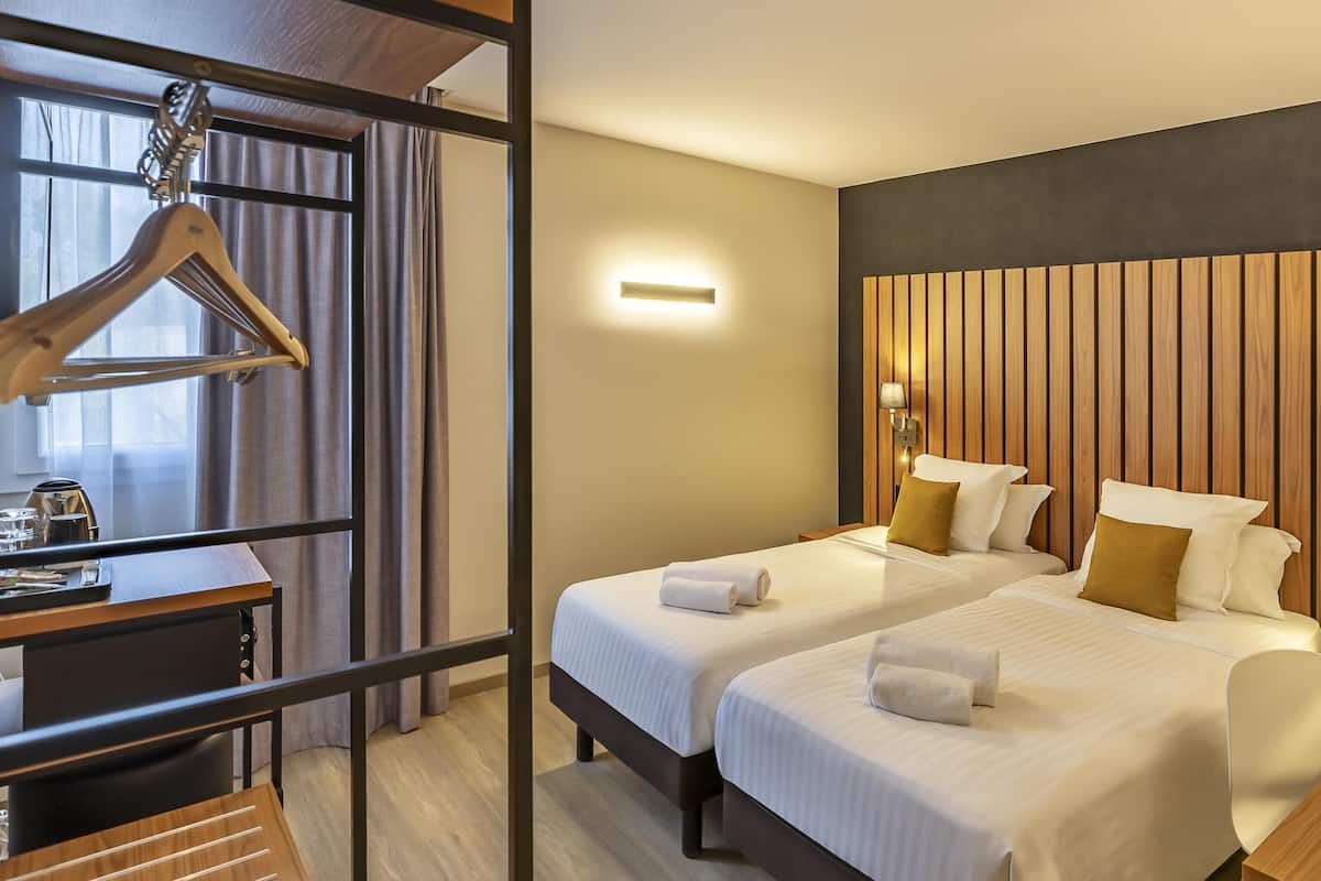 Une chambre d’hôtel avec deux lits, une tête de lit en bois, des lampes de chevet, un bureau avec une chaise et un porte-manteau mural.