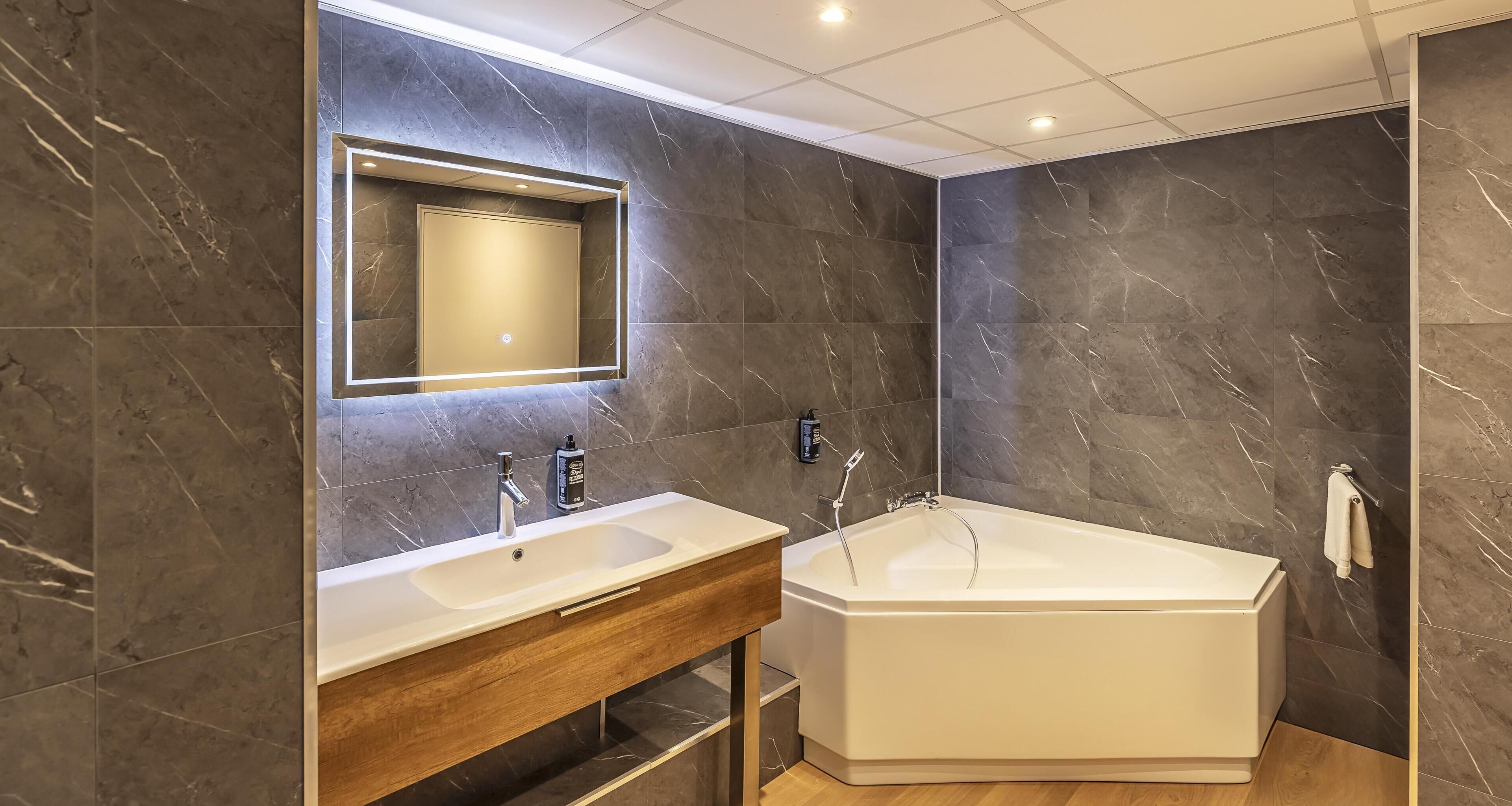 A modern bathroom with a marble wall, a large mirror, a white sink, a bathtub, and a wooden vanity.