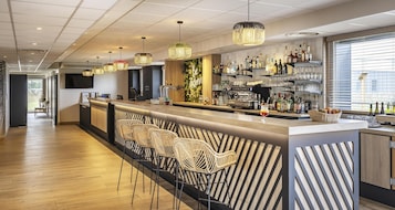 Un área de bar moderna con una barra larga, taburetes de mimbre y una variedad de botellas y vasos en estantes.