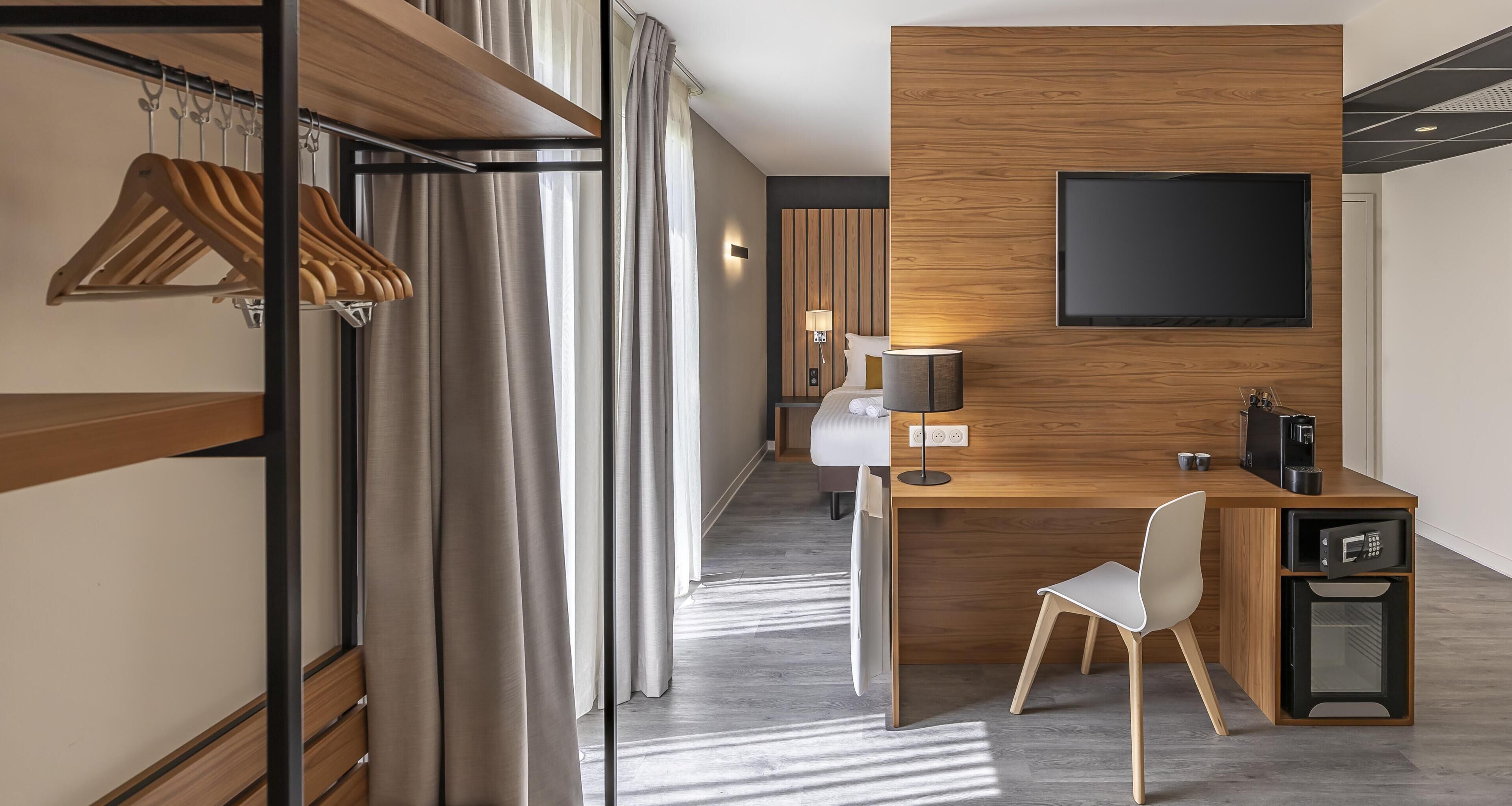 Een moderne hotelkamer met een houten bureau, een flatscreen televisie, een witte stoel, een magnetron en een kapstok met kleding.