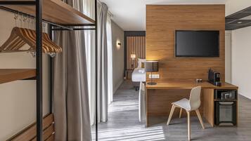 Une chambre d’hôtel moderne avec un bureau en bois, un téléviseur à écran plat, une chaise blanche, un four à micro-ondes et un porte-manteau avec des vêtements suspendus.