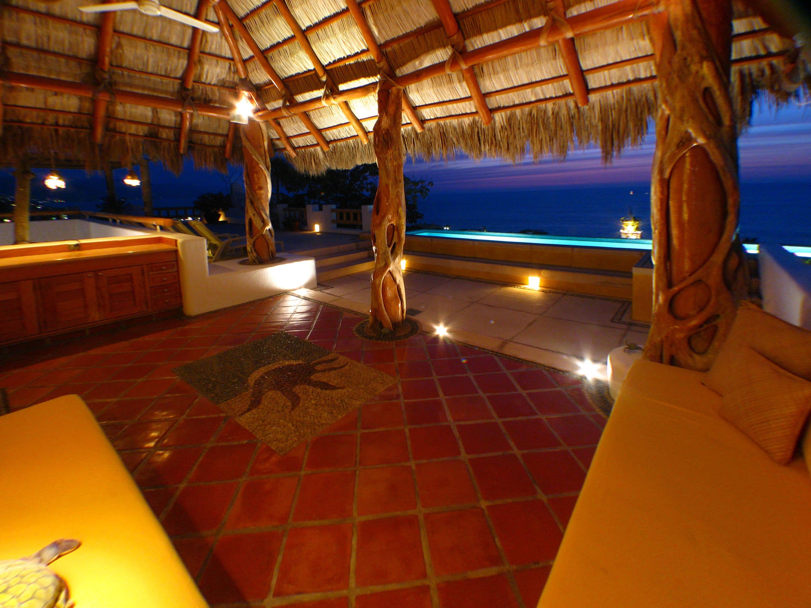 An outdoor lounge area with a thatched roof, tiled flooring, and a view of the sea at dusk.