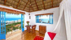 A room with a wooden floor, a bed with white and red bedding, a wooden cabinet, a round wooden table with a vase, and a window with a view of the ocean and buildings. - Luna Líquida Hotel Boutique (Puerto Vallarta)