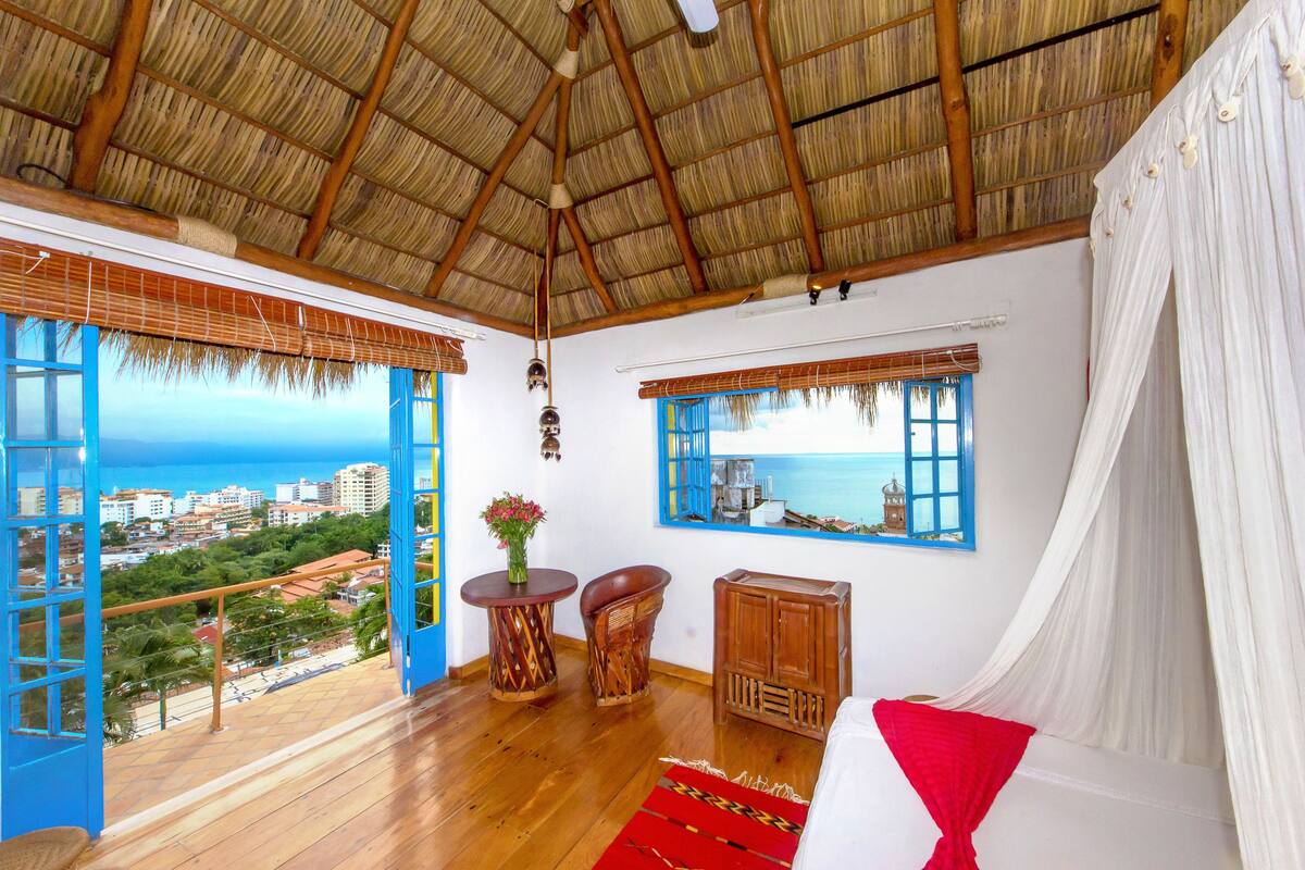 Ein Zimmer mit Holzboden, einem Bett mit weißer und roter Bettwäsche, einem Holzschrank, einem runden Holztisch mit einer Vase und einem Fenster mit Blick auf das Meer und Gebäude.