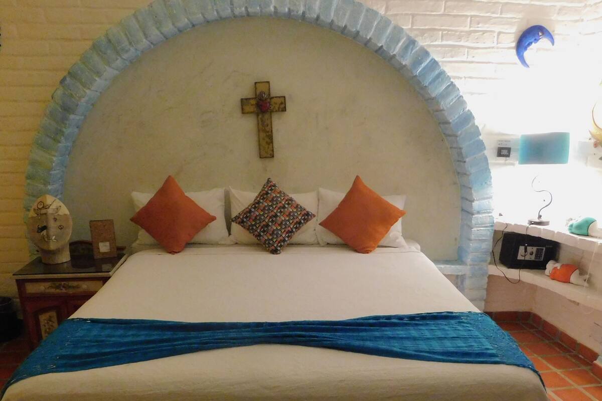 Ein Bett mit weißer Bettdecke, blauer Decke und drei Kissen. Über dem Bett befindet sich ein Kreuz an der Wand.