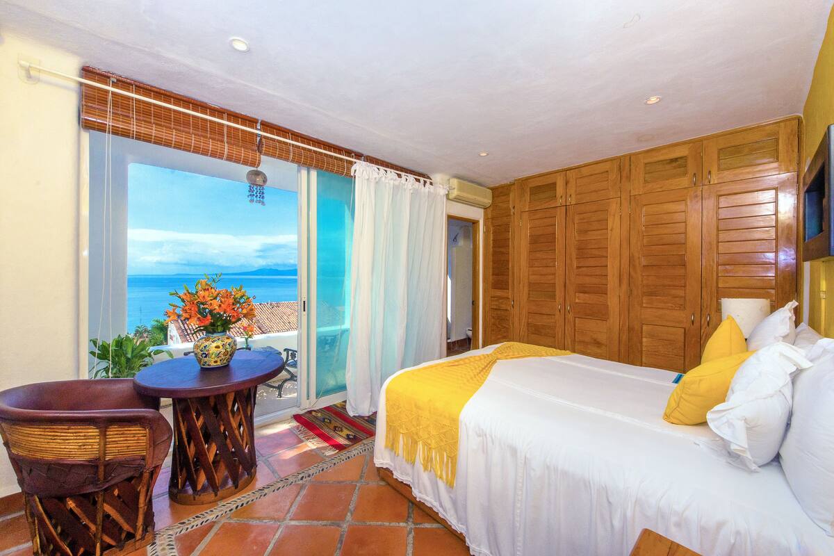 Ein Schlafzimmer mit Bett, Sessel, Tisch mit Blumenvase und Meerblick durch eine Schiebetür.