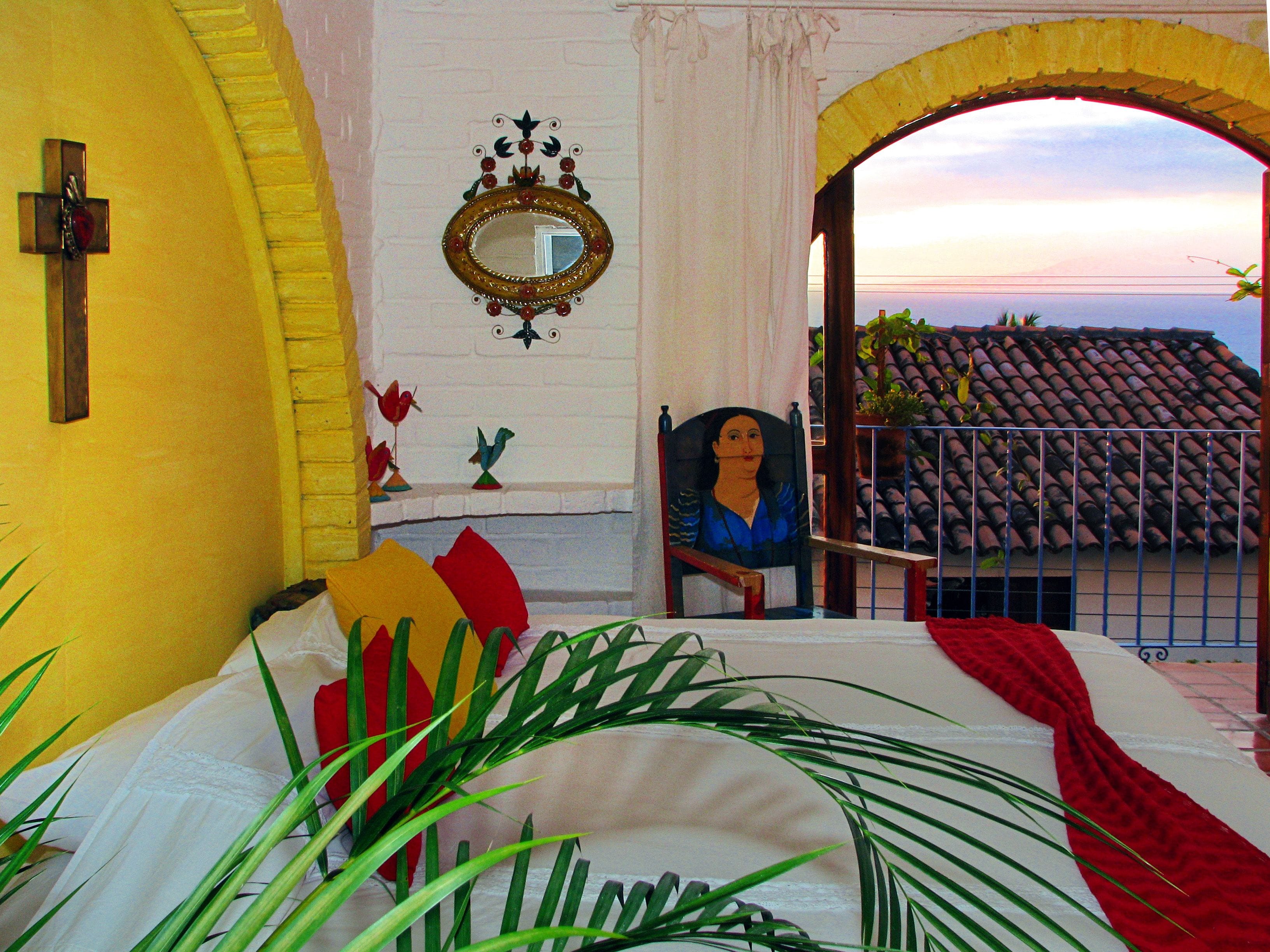 A room with a bed, a chair, a mirror, and a view of a tiled roof through an archway.