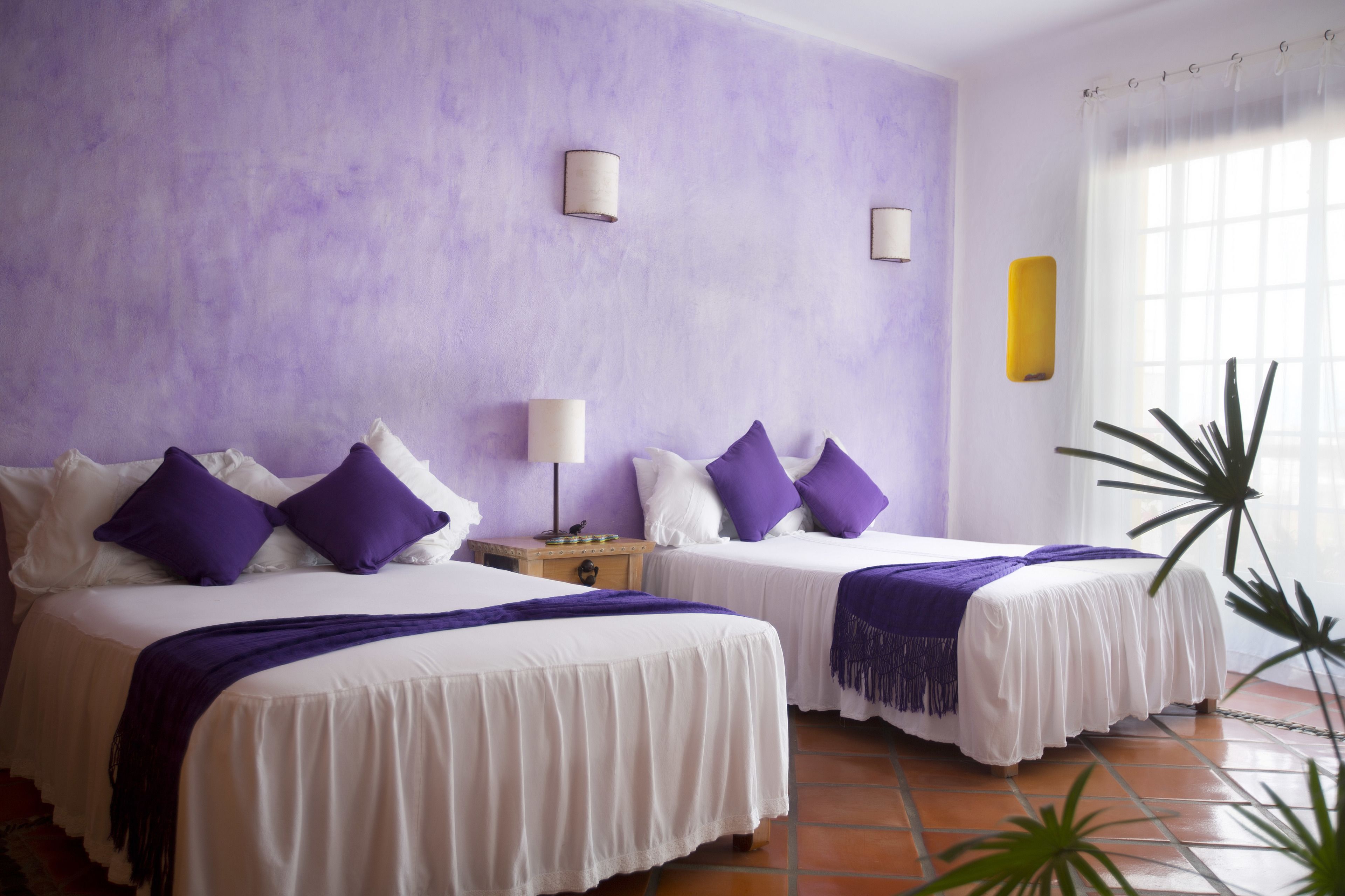 A hotel room with two beds, purple and white bedding, a nightstand, and a window with a curtain.