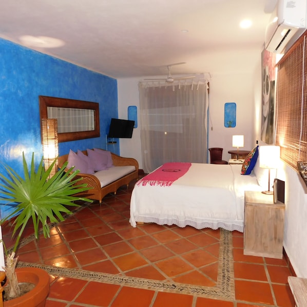 Habitación de hotel con una pared azul decorativa, cama con colcha blanca, sofá de madera, planta en maceta y piso de baldosas.