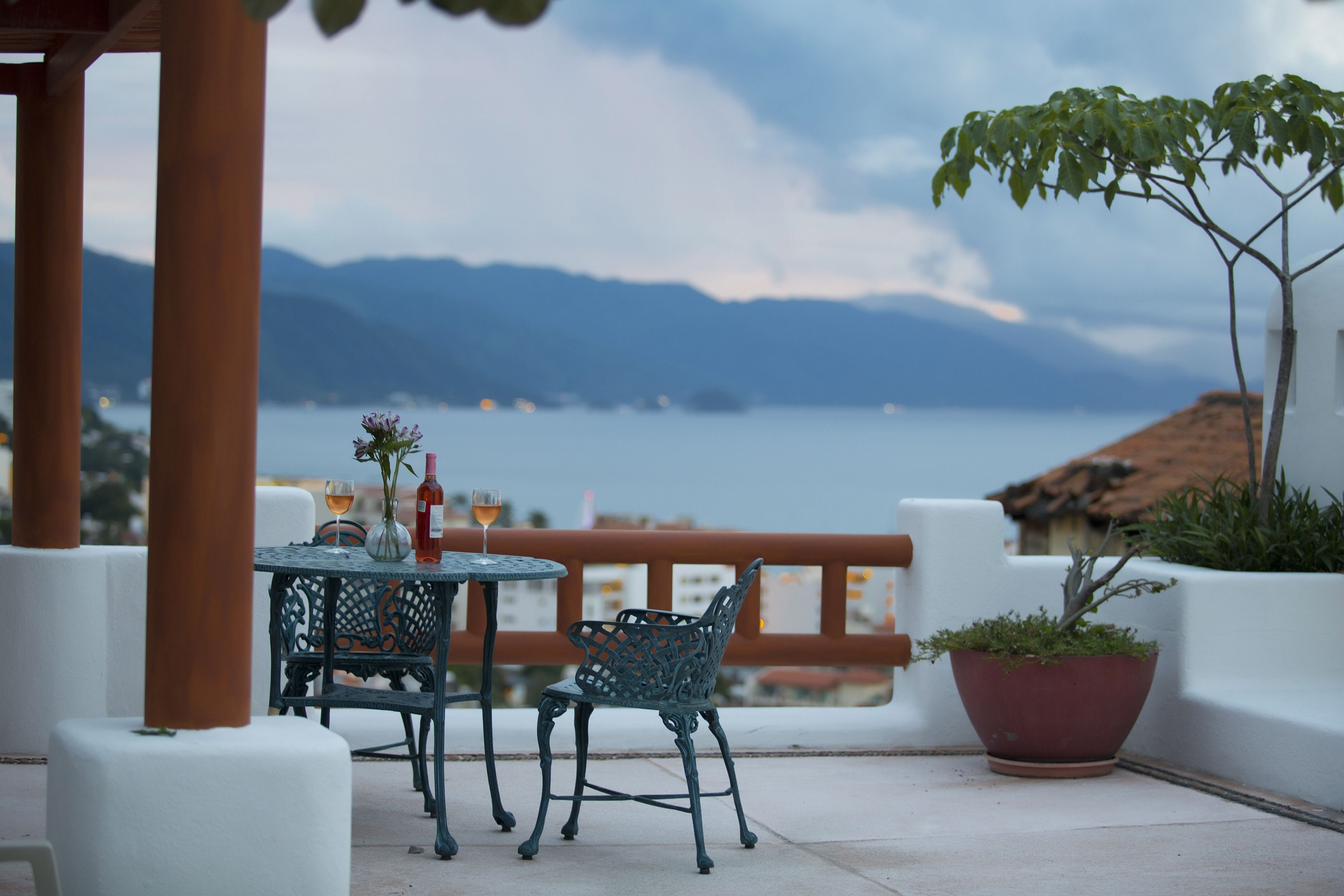 Un área de estar al aire libre con una mesa y sillas, una planta en maceta, y vista al mar y montañas al fondo.