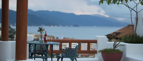 Un espace extérieur aménagé avec une table et des chaises, un pot de fleurs, et une vue sur la mer et les montagnes en arrière-plan.