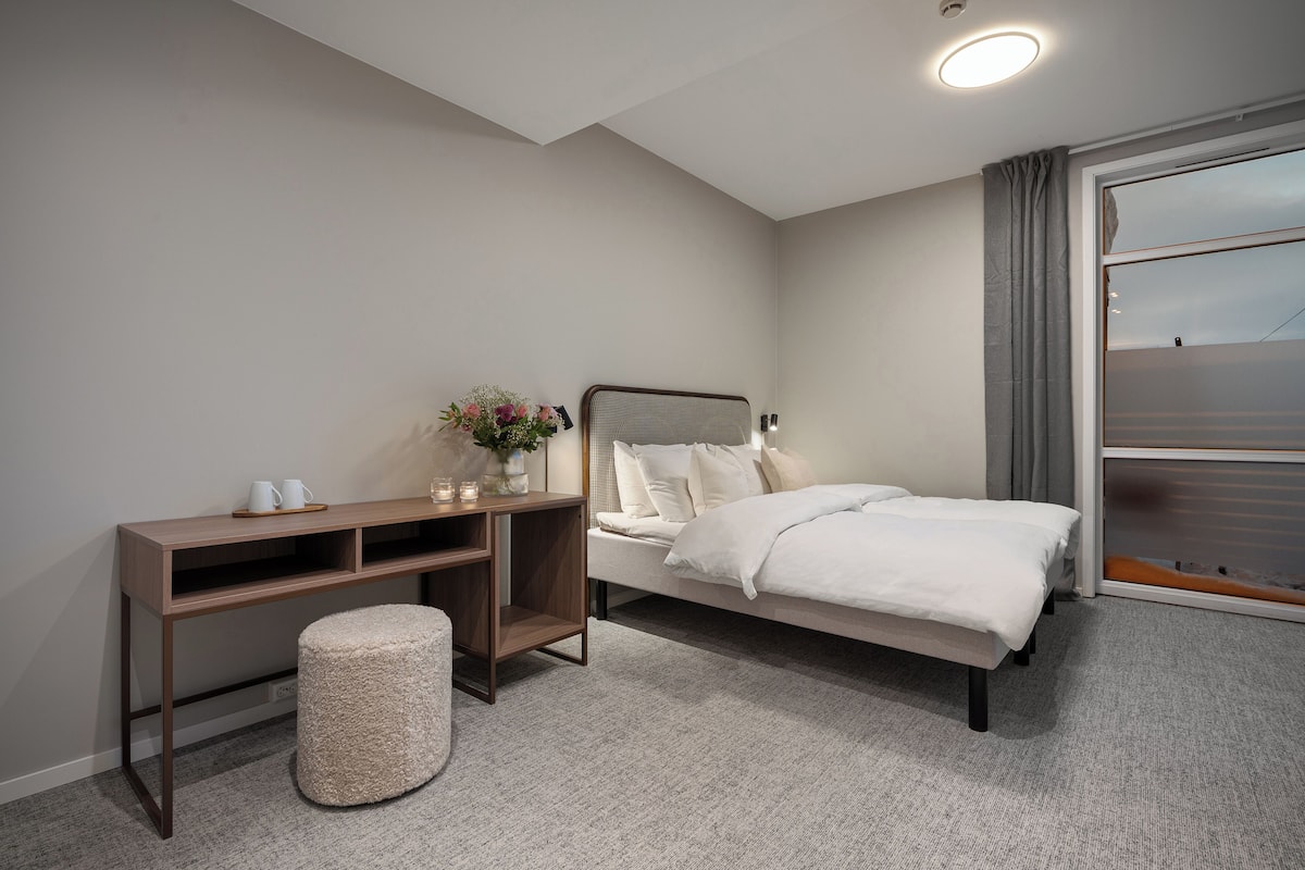 A bedroom with a bed, a bedside table with a lamp, a small stool, and a window with curtains.