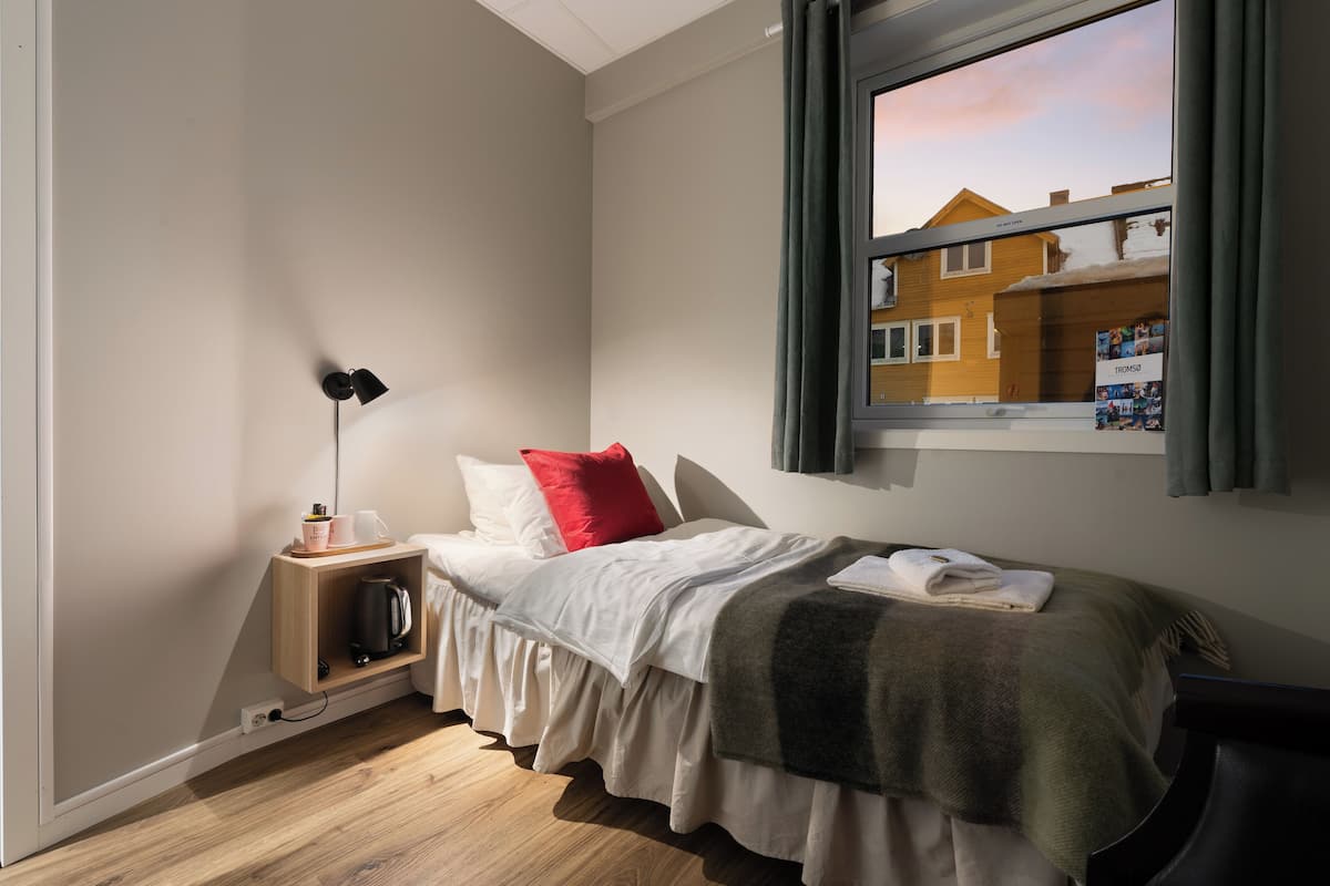 A bedroom with a bed, a bedside table with a lamp, a window with curtains, and a view of a house outside.