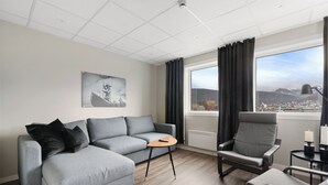 Ein modernes Wohnzimmer mit einem grauen Sofa, einem grauen Sessel und einem kleinen Holztisch. Auf einem Beistelltisch stehen eine schwarze Lampe und eine Kerze. Ein Schwarz-Weiß-Foto hängt an der Wand.