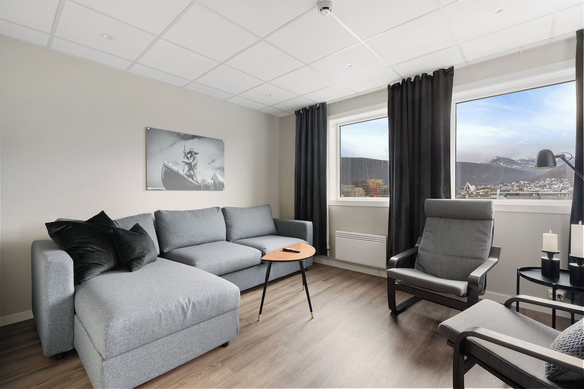 A modern living room with a grey sofa, a grey armchair, and a small wooden table. A black lamp and a candle are on a side table. A black and white photograph is on the wall.