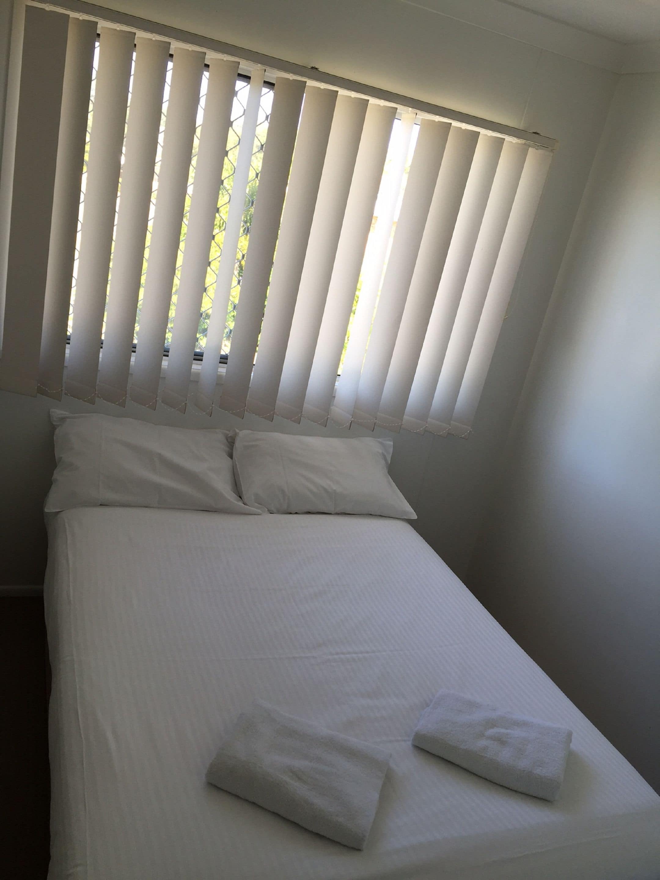 Un lit simple avec du linge de lit blanc et des oreillers, une fenêtre avec des stores verticaux et un mur uni.