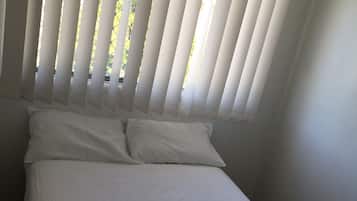 Un lit simple avec du linge de lit blanc et des oreillers, une fenêtre avec des stores verticaux et un mur uni.