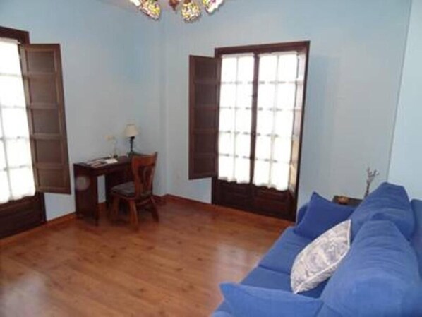 Una habitación con sofá azul, suelo de madera, un escritorio con silla y dos ventanas con cortinas.