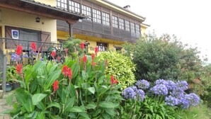 Un edificio de dos pisos con balcón, rodeado de exuberante follaje y flores coloridas.