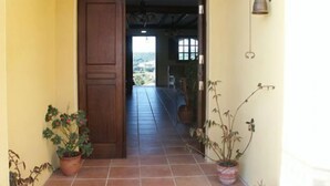 Un pasillo con una puerta de madera que da a una habitación, plantas en macetas y una lámpara fijada a la pared.