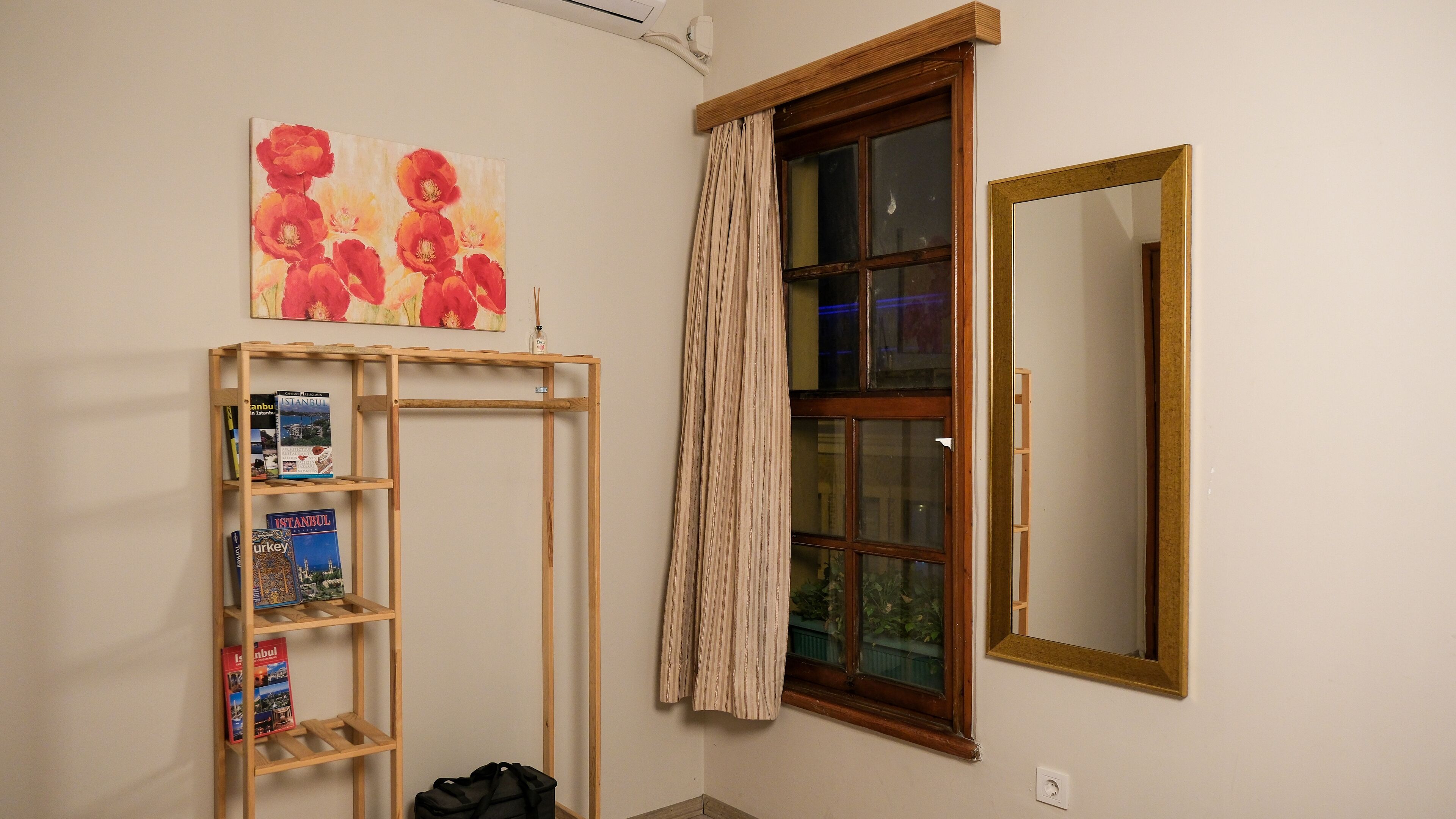 A room with a wooden shelf holding books and a painting, a window with curtains, and a mirror.