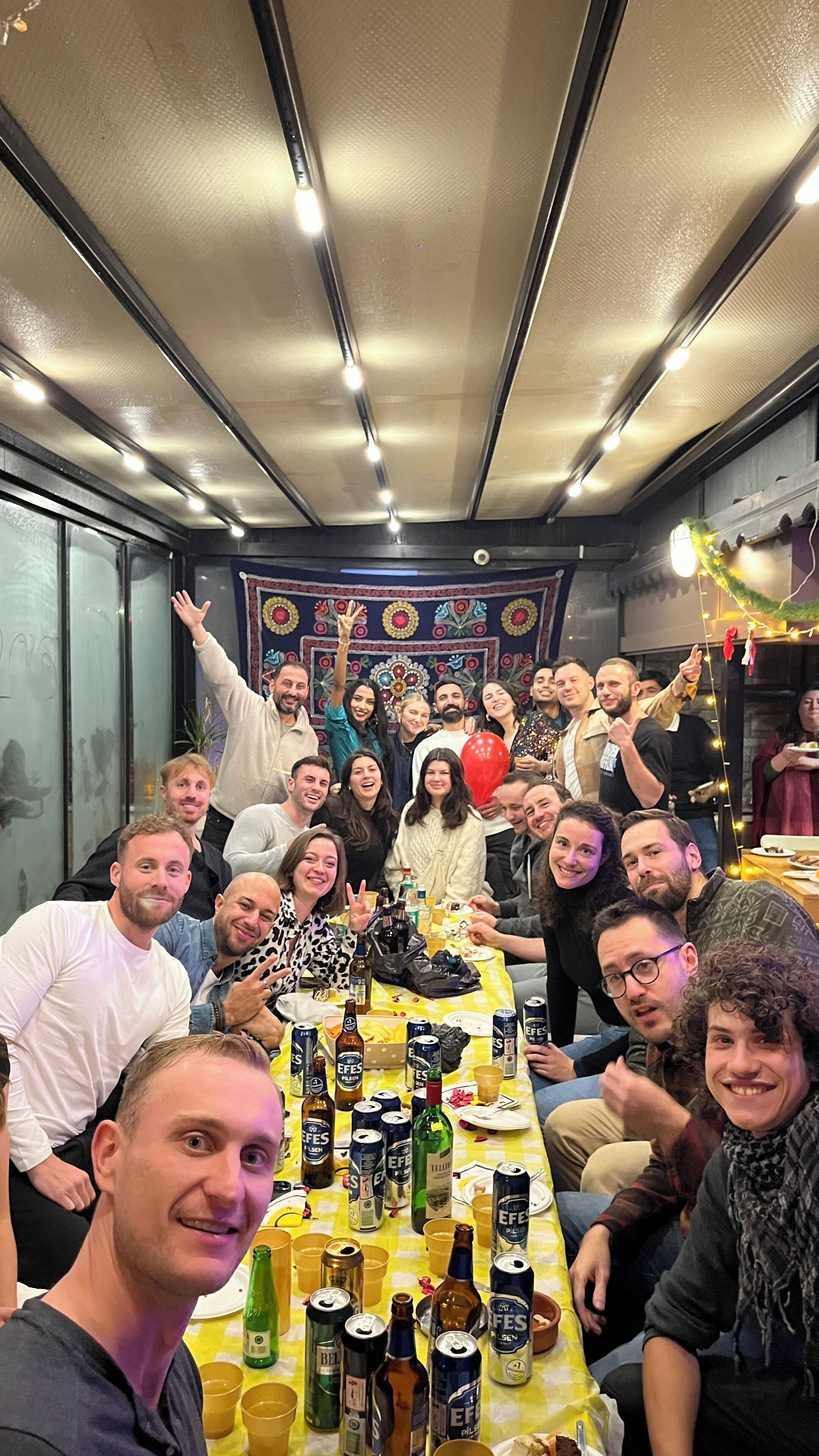 Um grupo de pessoas reunido em volta de uma mesa com vários frascos e latas, num ambiente com teto estampado e tapeçaria decorativa ao fundo.