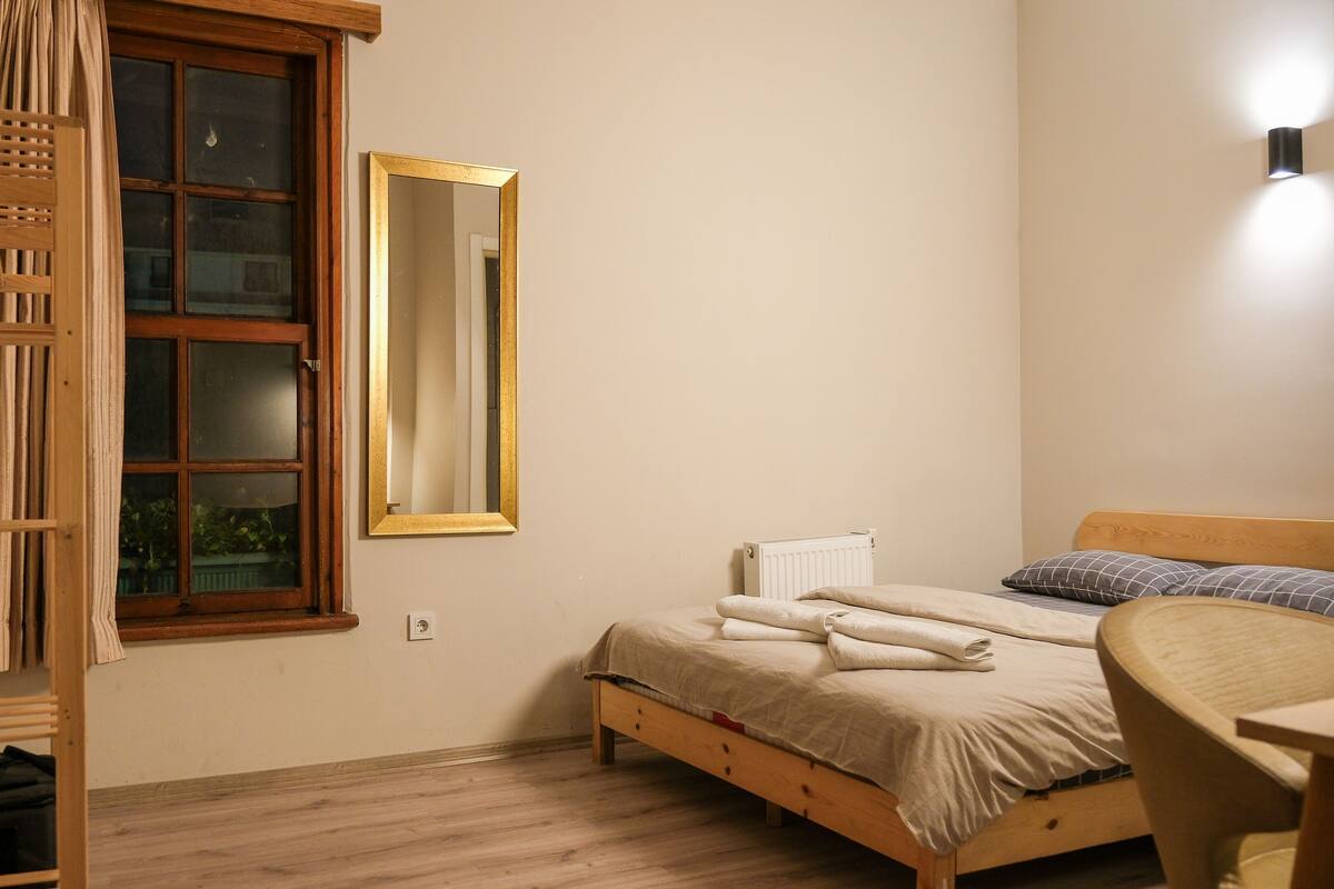 A bedroom with a wooden bed, a bedside table, a mirror, and a window with curtains.