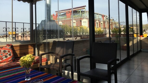 Une terrasse ensoleillée, dotée de chaises et d’une table en osier, offre une vue sur un bâtiment à l’architecture rouge et blanche.