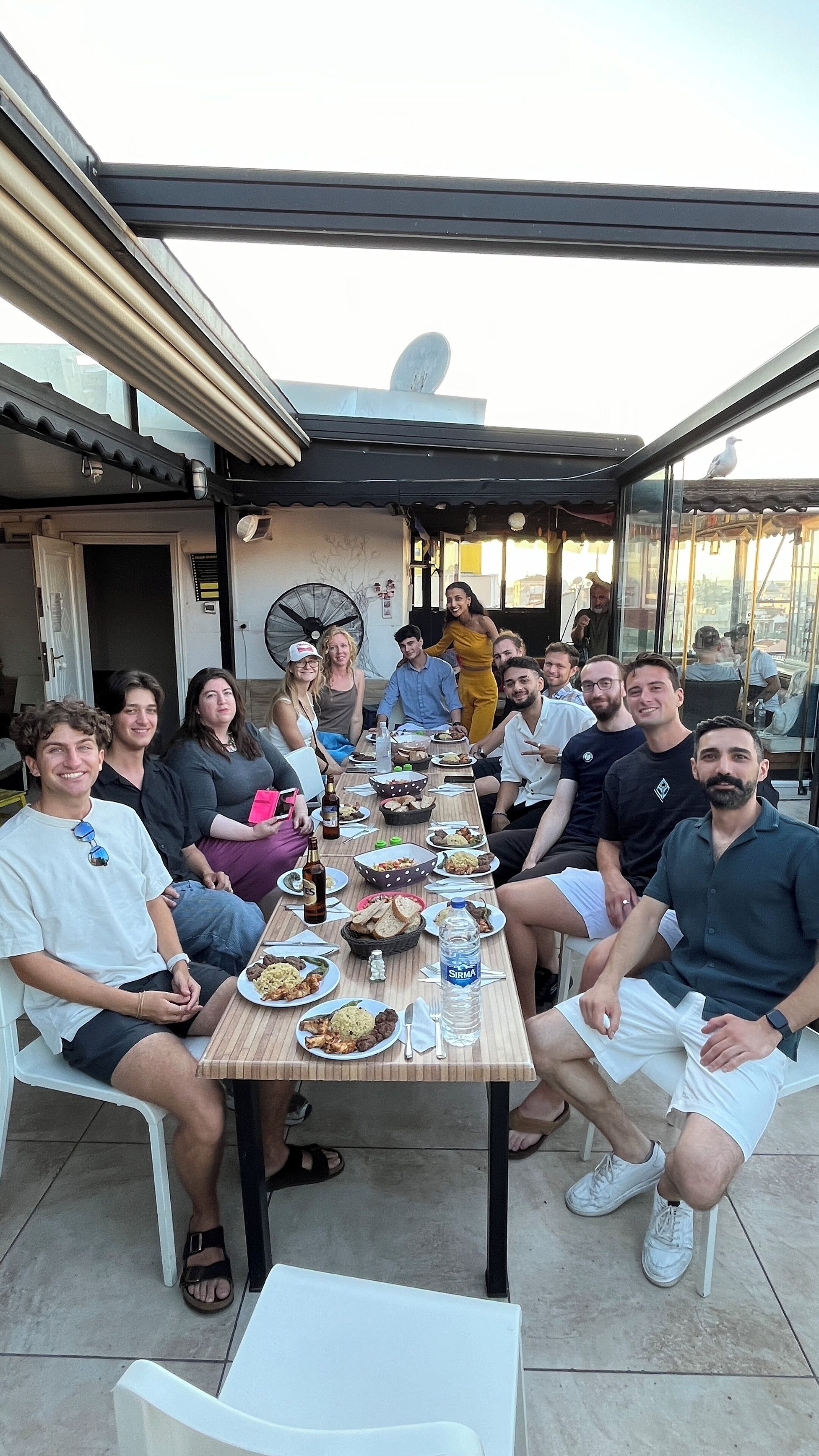 A group of people seated around a table with various dishes, in an outdoor setting with a covered patio.