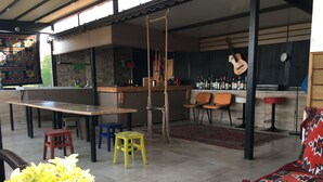 Un vaste espace de vie comprenant une longue table en bois, des tabourets colorés, une guitare suspendue et un coin bar avec des bouteilles.