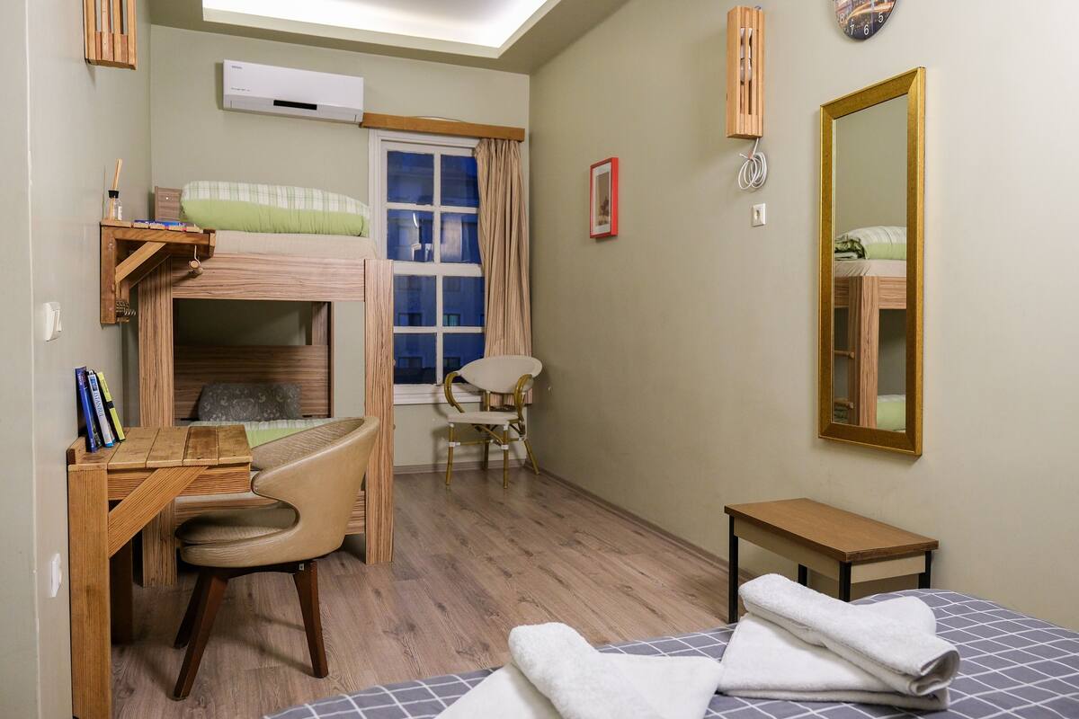 A compact room with a bunk bed, a desk, a chair, a small table, and a mirror.
