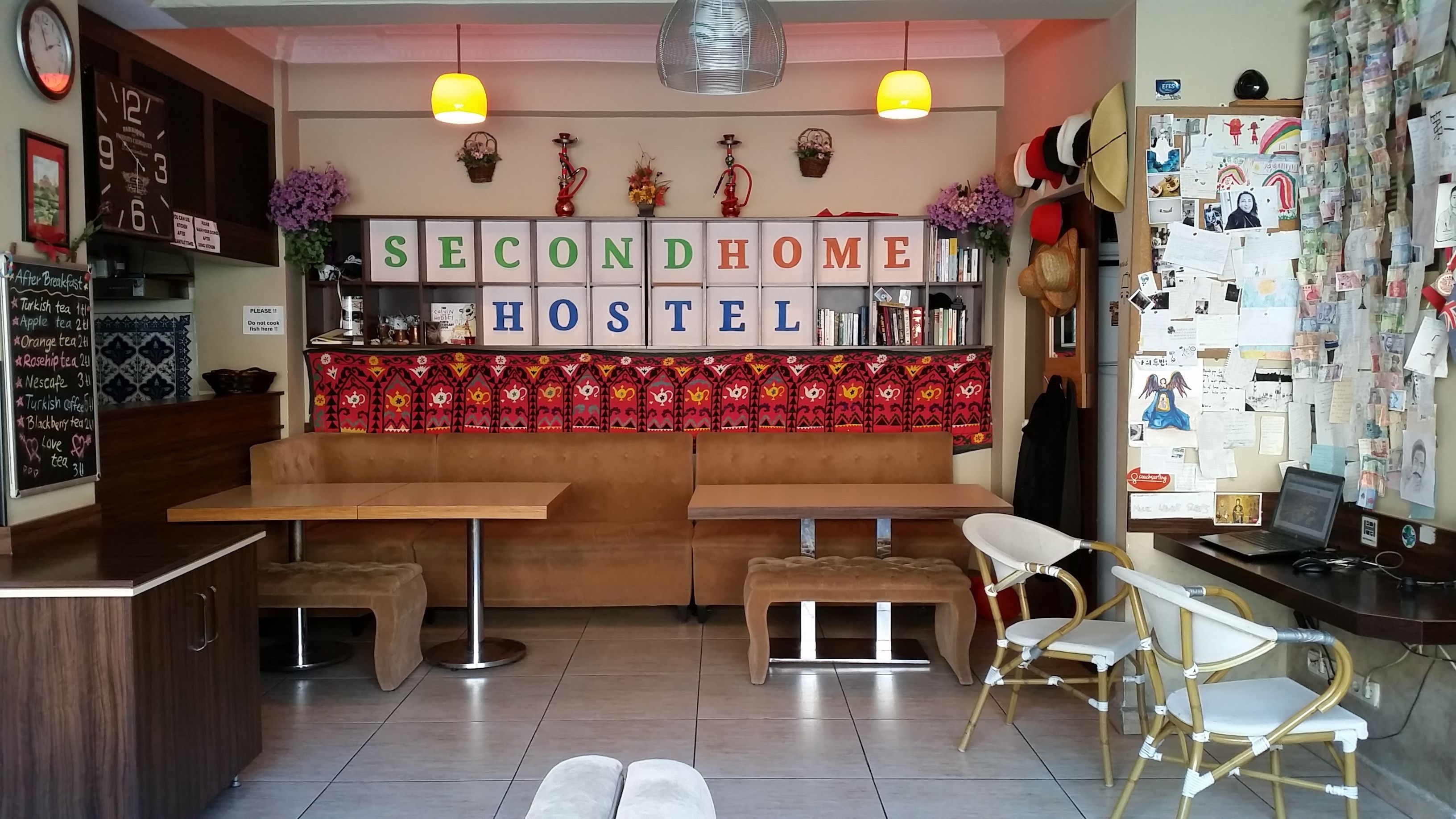 A cozy hostel with a central seating area, a bar, and a wall decorated with various items including a sign that reads "SECONDHOME HOSTEL".