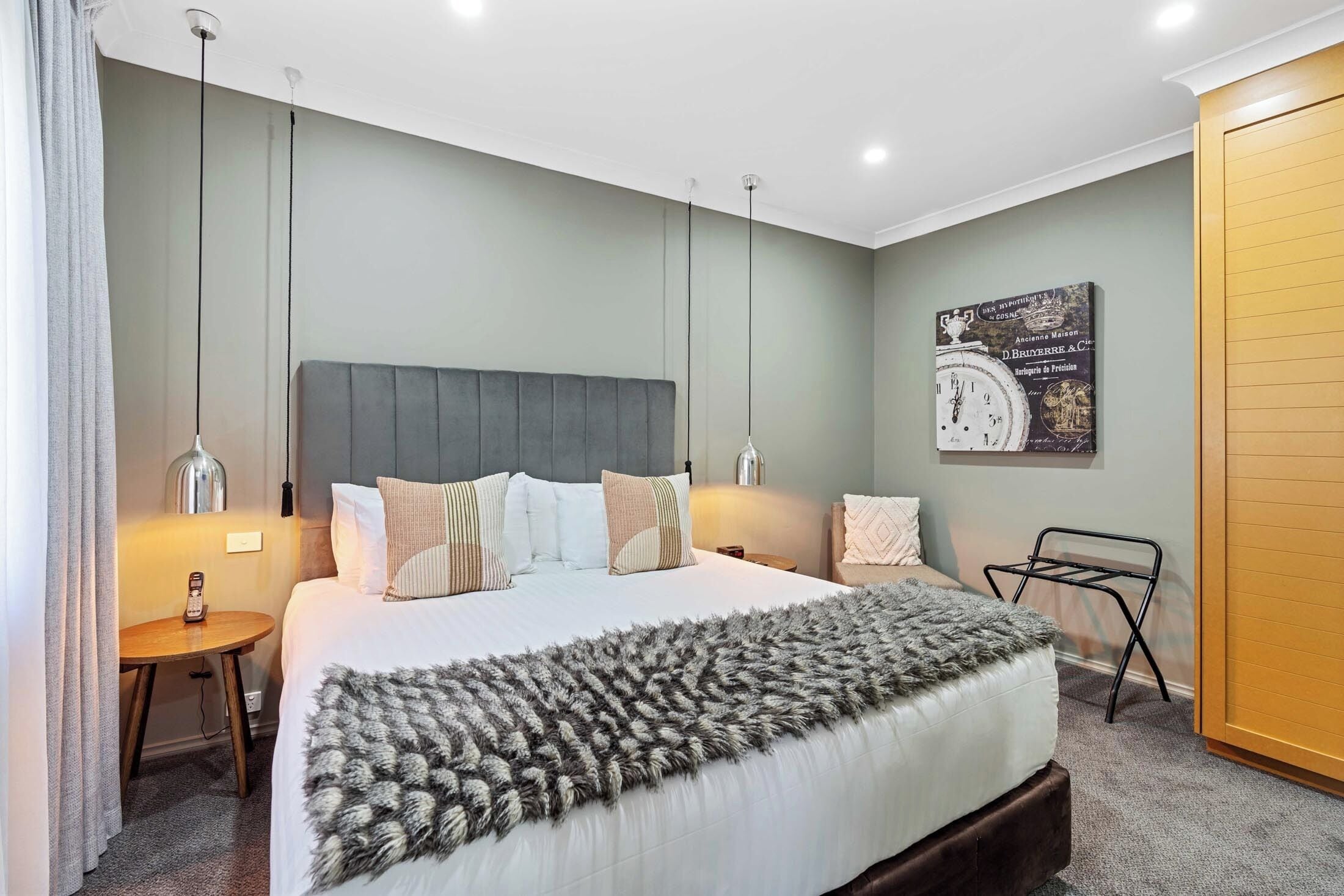 Une chambre à coucher bien rangée, avec un grand lit, deux tables de chevet, une horloge murale et une petite table d’appoint.